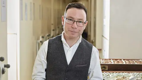 Douglas Russell A smiling Douglas Russell looking directly at the camera, wearing a white shirt and dark waistcoat