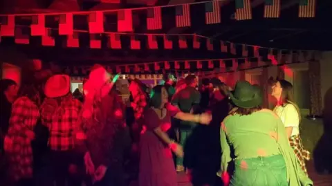 Perry St Social Club People dressed in cowboy outfits dancing under red lights and USA flag bunting in a bar