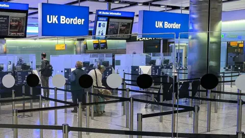 People queue at border control in a UK airport, with a sign above reading "UK Border".