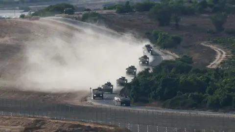 An Israeli military convoy manoeuvres near the Israel-Gaza border. Armoured vehicles can be seen driving along a winding road, with greenery to the right and sand to the left. A fence can be seen in the foreground.