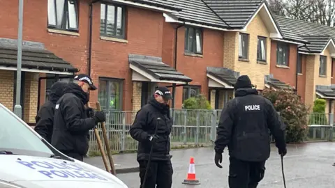 Four police officers, all wearing black with baseball caps, standing on a street, with several houses behind them