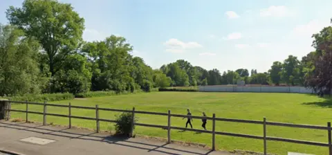 A large green field with a border of trees and bushes. two people walk over the grass. in the foreground is wooden fencing slightly higher than the field. on the other side of it is a concrete path and a road 