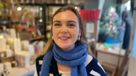 Meghan Kinnison-Cook standing inside Domestic Science. She is a young woman wearing a blue wool scarf and blue and white jumper. She has brown hair and is smiling at the camera. She's got big silver hoop earrings on. 