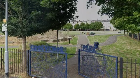 Google The entrance to Grosvenor Park, signposted as such above some blue gates. Trees, a footpath and grassed areas can be seen in the background.