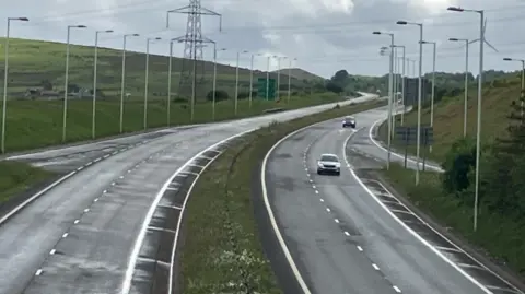 A general view of the A465 dual carriageway, which has two lanes travelling in each direction separated by a grass verge. Lampposts line each side of the road, with an electricity pylon on the left hand side and a hill beyond.
