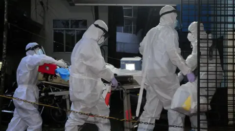 Four medical workers in white protective clothing move an unseen patient on a hospital trolley.