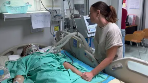 A photo of someone in a hospital bed lying down and wearing a green hospital outfit. They have got breathing machines attached to them and a woman with brown hair is standing and holding his hand. 