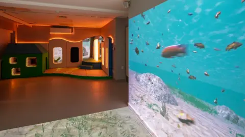 Martin House/JDDK Architects/Meldrum Group An indoor children's play area which is lit up by LED lighting behind a soft play area. There is an undersea image with fish and the sea floor projected onto one of the walls and flooring.