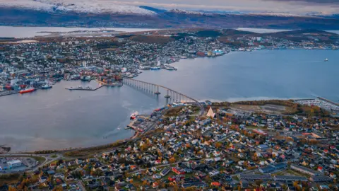 Getty Images An aerial view of Tromsø, Norway, showing the Tromsø Bridge spanning the water to connect the city. Densely packed buildings and harbours sit below a backdrop of snow-dusted mountains under a pastel-coloured sunset sky.