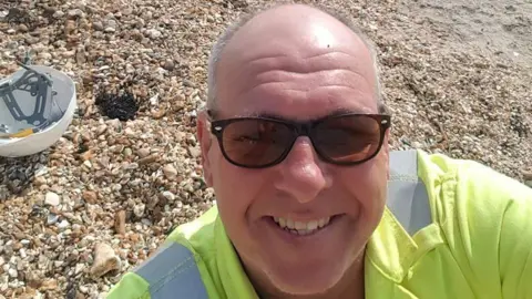 Steve Dymond/Facebook Steve Dymond looks at the camera in a selfie photo. There is a shingle beach in the background and a builder's hardhat on the ground