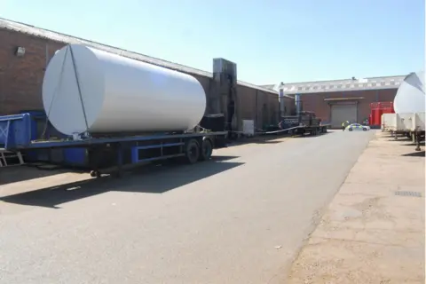 HSE An exterior view of an industrial building, with one entrance shown in the distance. A large white cylinder on the back of a flatbed trailer is in front of the building.