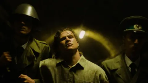 An image taken from the short film Whispers of Freedom. Set in East Germany in the 1980s it shows two men in officers uniforms accompanying a man in a checked shirt.

