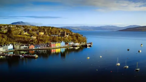 Getty Images Sebuah pemandangan udara Tobermory, dan rumah -rumahnya yang berwarna cerah, dan teluknya dengan kapal pesiar berlabuh di dalamnya. Ada bukit -bukit di pantai di latar belakang.