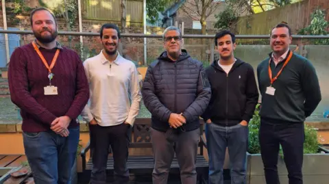 Five men are standing in a row and posing for an image. They appear to be standing outside and behind them is a metal railing and a garden with trees. They are wearing jumpers and coats.