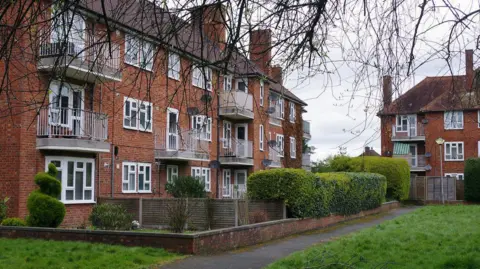 Steve McKay/Geograph Three-storey brick-built apartment buildings with balconies on the 1st and 2nd floor windows. There is a low brick wall around the block, and there are hedges in front of some of the apartments. A further block is visible in the background, and there is a grassed area to the right, fringed by a path.