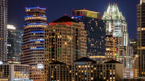 Getty Images Night time city skyline with illuminated high-rise buildings and office towers, featuring distinctive modern architecture with lit windows and colourful accent lighting against a dark sky.