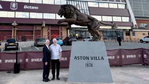 Tanya Russell Two men standing next to a large, bronze lion statue behind the North Stand at Villa Park.