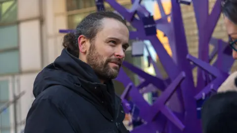An image showing the side-profile of a man with dark hair in a ponytail, wearing a black coat and standing next to a purple tree-like sculpture.