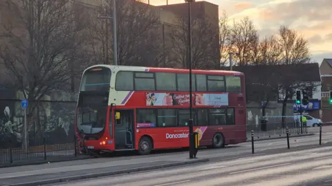 A double-decker bus at a standstill after crashing into some black railings, with police tape in the background.