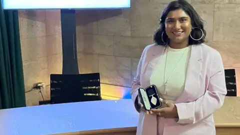 ZeZe Sohawon A woman wearing a light pink blazer holds a medal in a box in her hands.