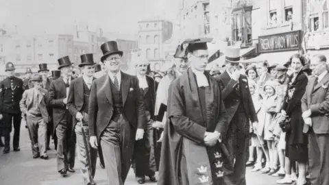 Bettmann/Getty Images A black and white image showing the centre of Boston, UK. A civic party is processing through the town. The men are wearing morning suits and top hats. Joseph Kennedy is pictured in the first row of the procession. Crowds stand to the side looking on.