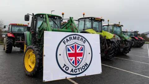 Tractors are parked up at the Cornwall Services. The one nearest to tthe camera has a large sign reading FARMERS TO ACTION with a shield formed of the Union Jack flag tied to its bucket.