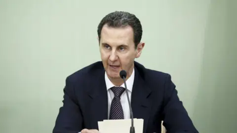 GettyImages Bashar al-Assad pictured in 2023 speaking into a microphone and holding some papers. He is wearing a dark suit, white shirt and tie.