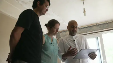 Couple Freya and Alex standing in a room looing at a tablet held by architect Imran