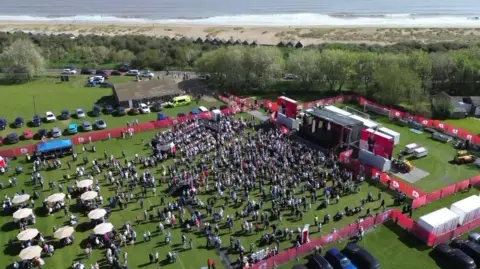 An aerial shot of the event - a football pitch with a large stage, crowds of people, and picnic tables with parasols.