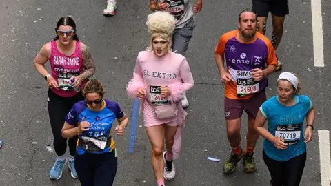 Getty Marathon runners move along a city street, including a participant wearing a pink costume, as others run alongside.