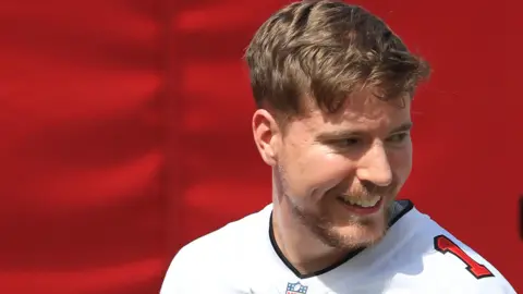 Getty Images Jimmy Donaldson, também conhecido como Sr. Fera, tem cabelo castanho curto e barba curta. Ele usa um uniforme branco de futebol americano com um número vermelho no ombro esquerdo. Ele olha para a câmera por cima do ombro esquerdo e é fotografado contra um fundo vermelho. 