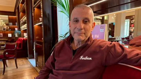 A middle-aged man in a burgundy quarter-zip fleece looks directly into the camera with a subtle smile. Behind him is a restaurant 