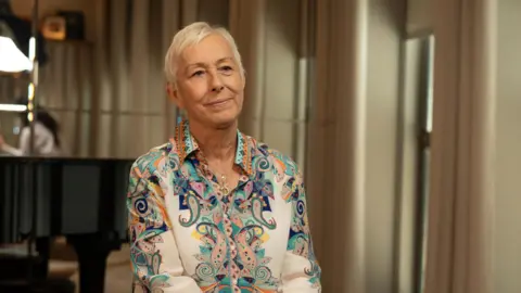 Martina Navratilova, with short white hair, wearing a colourful paisley-print blue and orange collared shirt and a thin gold chain necklace. She's sitting in a room in front of some beige curtains - the background of the photo is out of focus.