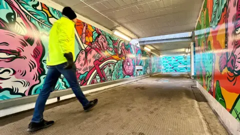 A man in a bright yellow high-vis jacket, black bobble hat and blue jeans walks through an underpass at Bedminster Station in Bristol. All the walls around him have colourful cartoon-style murals on them