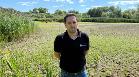 Karl Curtis wearing a black polo shirt with a Warwickshire Wildlife Trust logo, stands in front of a dried out lake bed at Brandon Marsh 