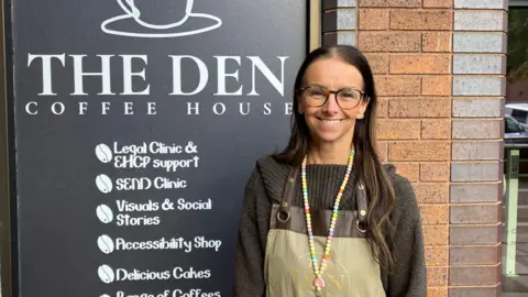 Claire Naylor is wearing a black jumper with an apron, and glasses, standing in front of a large sign for The Den Coffee House which lists its services. Services include a Send clinic, an accessibility shop and support with education, health and care plans.