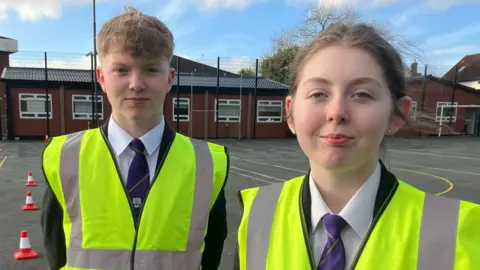 Matthew Blair (left) and Amy McCoy (right). Matthew has strawberry blonde hair, Amy has brown hair tied in a ponytail. Both are wearing their school uniform - black blazer, white shirt and blue tie - and high viz vests. They are in the school playground with the school building in the background.