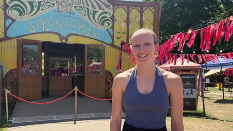 James McConnell/BBC Chelsea Hall stands in front of the Norfolk and Norwich Festival's yellow circus tent on a sunny day. She is smiling and wearing a purple halterneck leotard. Her hair is blonde and pulled back from her face.