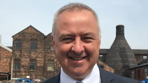 Ben Adams is in Stoke-on-Trent standing in front of a bottle kiln, smiling at the camera.
