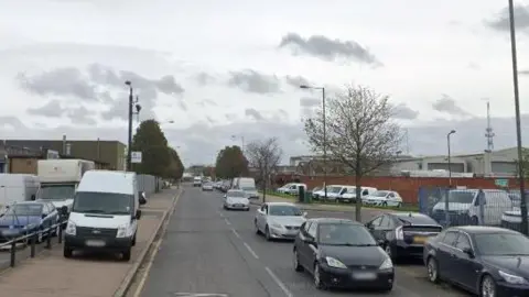 A general view of Nathan Way in Thamesmead. There are cars and vans parked on either side of the road. 