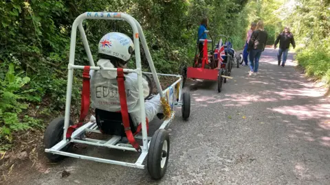 A narrow road with two soapbox carts parked on the lefthand side. People walk along the road, which has woodland shrubs and trees growing either side of it.