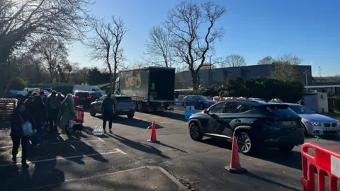 A number of people milling around the Tunbridge Wells Sports Centre on St John's Road, Tunbridge Wells. Cars can be seen waiting in a queue for water.