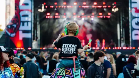 A crowd at Teddy Rocks festival in front of their stage. A child is seen on the shoulders of an adult in the middle of the crowd. The child is wearing a black shift reading Teddy Rocks 2025.