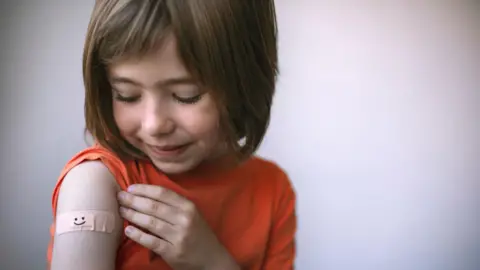 A young girl with brown, shoulder-length hair has rolled up the sleeve of her orange t-shirt to reveal a plaster with a smiley face on it on her upper arm, where an injection might go.