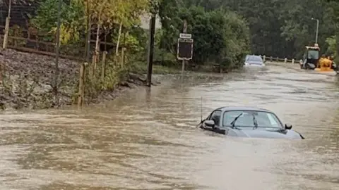 A badly flooded road engulfs a black car while a tractor can be seen in the background.