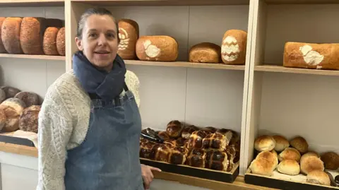Amy Holmes/BBC A woman wearing an apron stands in a bakery in front of shelves filled with various freshly baked loaves and rolls.