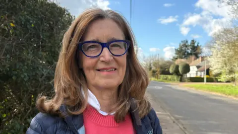 Sue Clark with long brown hair and blue-framed glasses smiling at the camera while wearing a pink sweater and purple puffer jacket. She is standing next to a rural road with a white country house in the background.