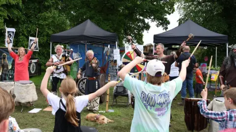 Supplied In a field, the Biking Vikings, dressed as Vikings, are performing in a line playing various instruments including the pipes, drums and strings. There are children facing them, holding drumsticks, with their arms raised too. 