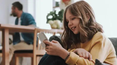 Young girl in yellow jumper stares at a mobile phone. her dad can be seen in the background