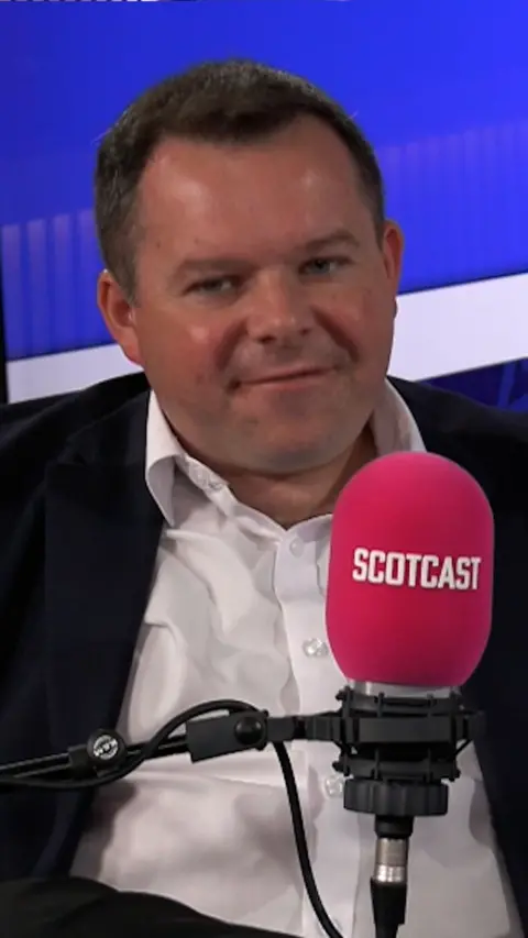 Man in white shirt and black jacket in radio studio with blue background.